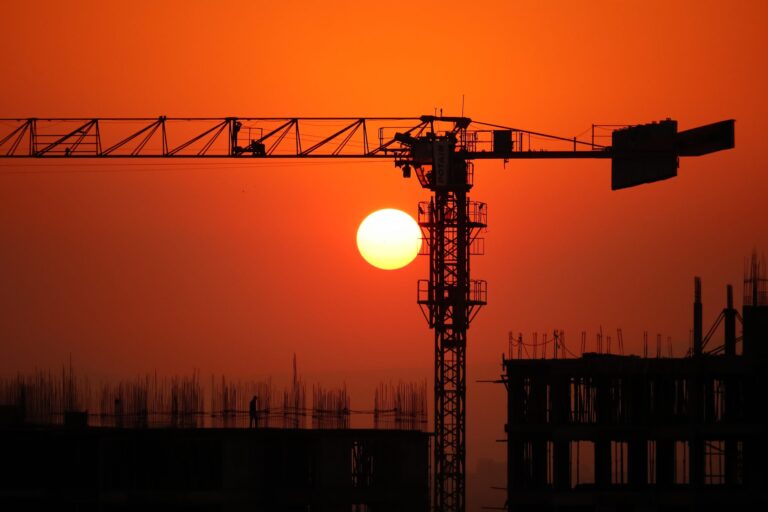 Sun setting in an orange sky with a silhouetted crane hovering over construction