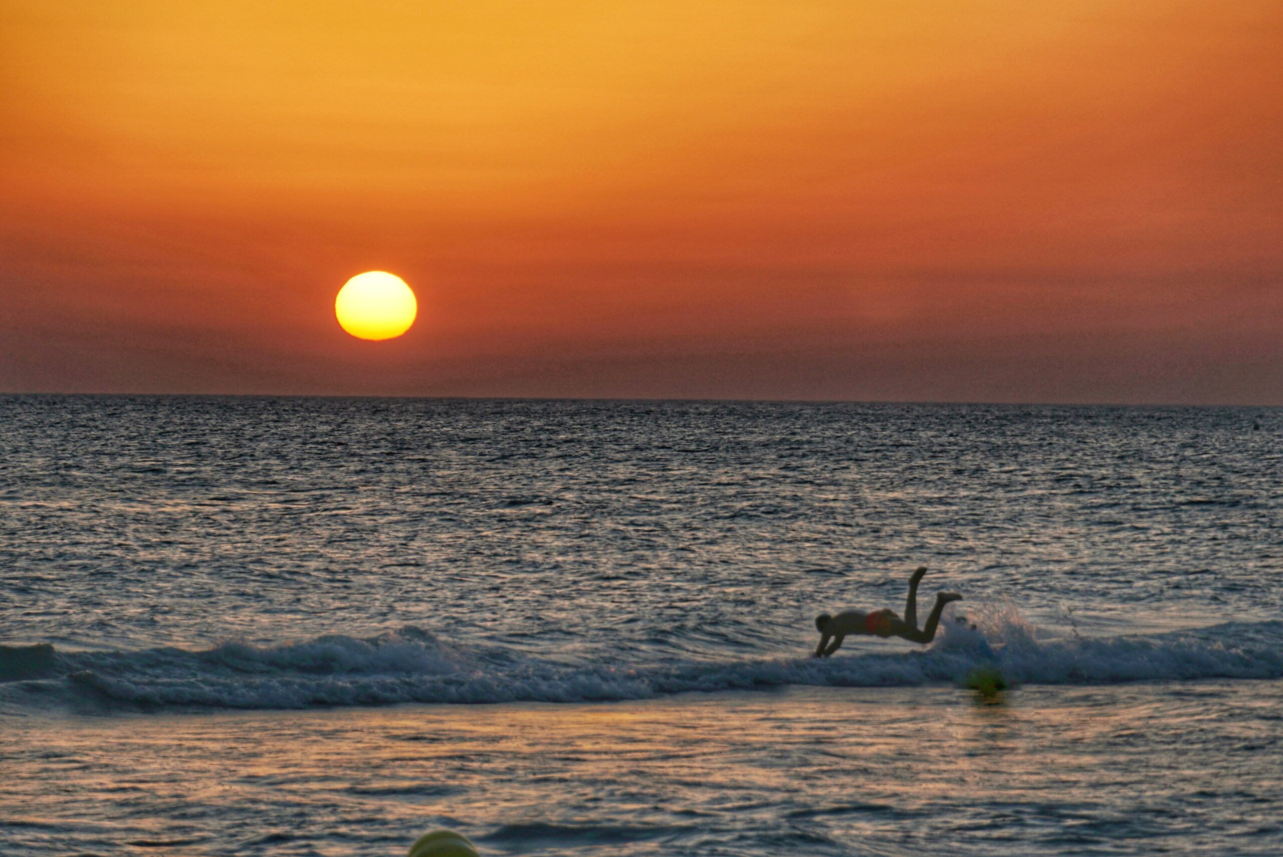 The sun setting over the ocean with a person diving into the surf.